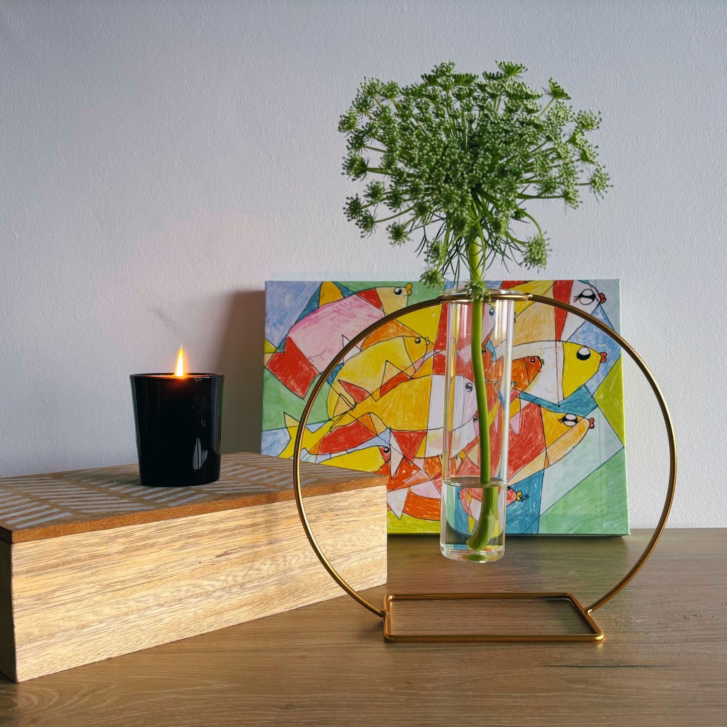Decorative setup with a vase, candle, and abstract painting on a wooden surface.