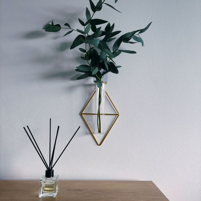 Decorative diffuser with black reeds and a geometric wall vase with greenery on a light gray background.