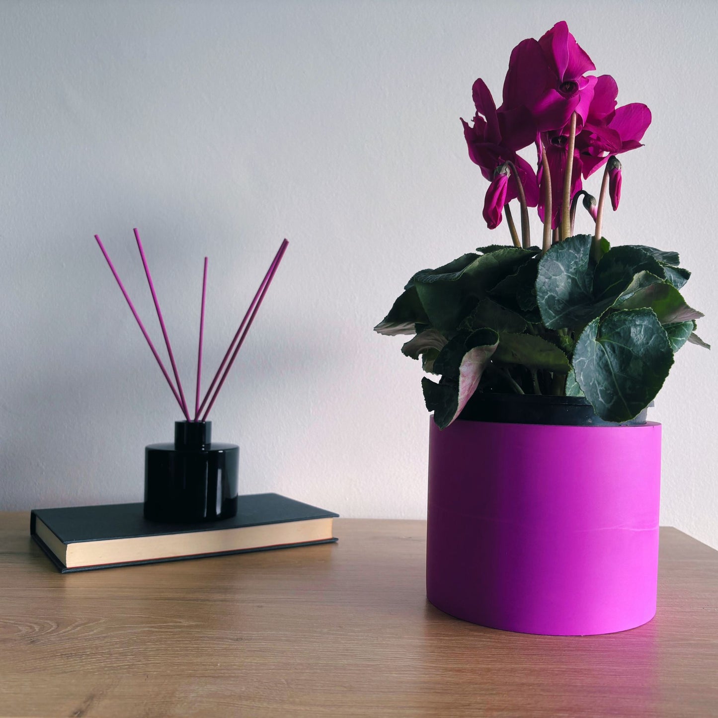 Purple plant pot with a pink flower on a wooden surface, next to a black book and diffuser.