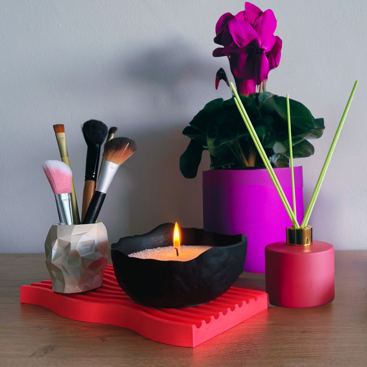 Makeup brushes in a holder, lit candle, purple plant, and pink diffuser on a wooden surface.