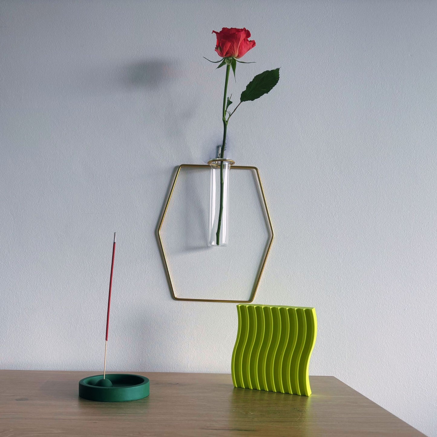 Green wavy vase with a red rose and incense stick on a wooden surface against a white wall.