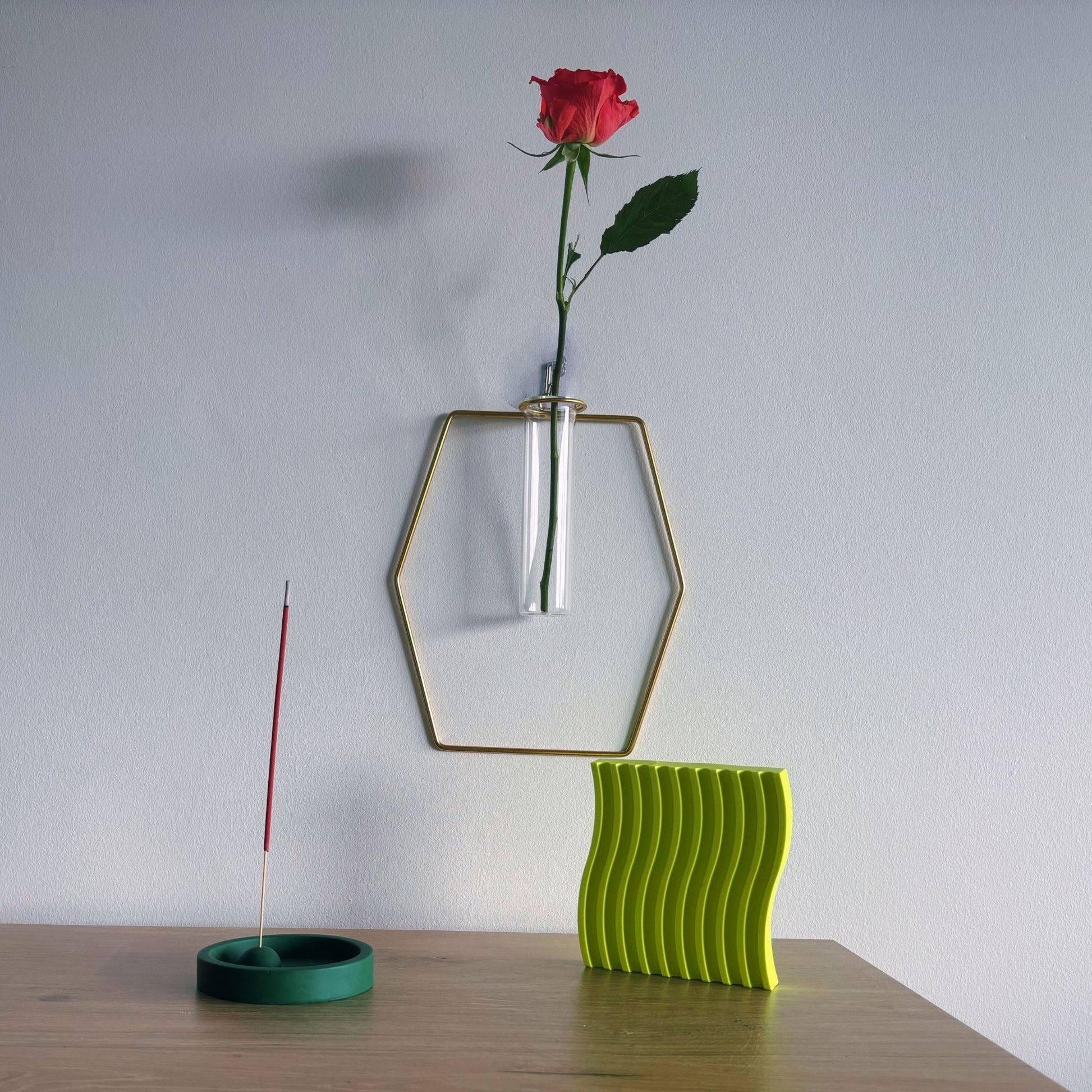 Green wavy vase with a red rose and incense stick on a wooden surface against a white wall.
